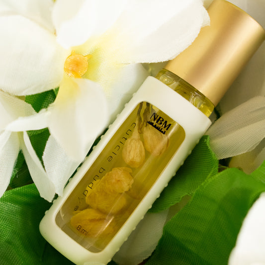 a bottle of cuticle oil with a gold cap, white silicone holder, and jasmine buds inside next to a faux jasmine flower and on top of faux green leaves