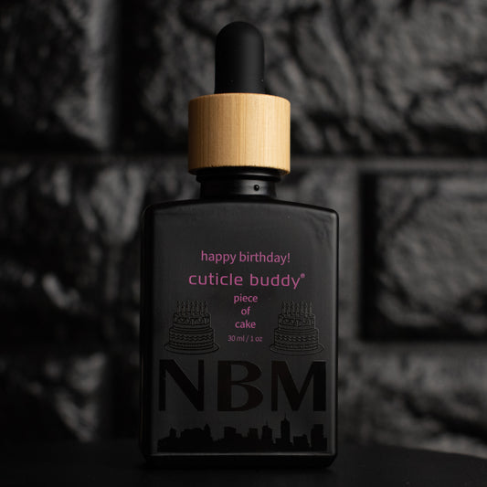 matte Black bottle with wooden dropper labeled  happy birthday! cuticle budd piece of cake 30ml/1 oz with cake graphics and a big NBM logo on the bottom featuring a city skyline. on a dark background