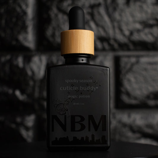 matte Black bottle with wooden dropper labeled  spooky season cuticle buddy magic potion 30ml/1oz with a magic potion graphic and a big NBM logo on the bottom featuring a city skyline. on a dark background