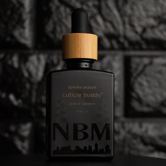 matte Black bottle with wooden dropper labeled  spooky season cuticle buddy  jack-o'-lantern 30ml/1 oz with jack o lantern graphics and a big NBM logo on the bottom featuring a city skyline. on a dark background