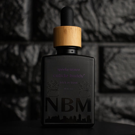 matte Black bottle with wooden dropper labeled  spooky season cuticle buddy trick or treat 30ml/1 oz with bags of candy graphics and a big NBM logo on the bottom featuring a city skyline. on a dark background