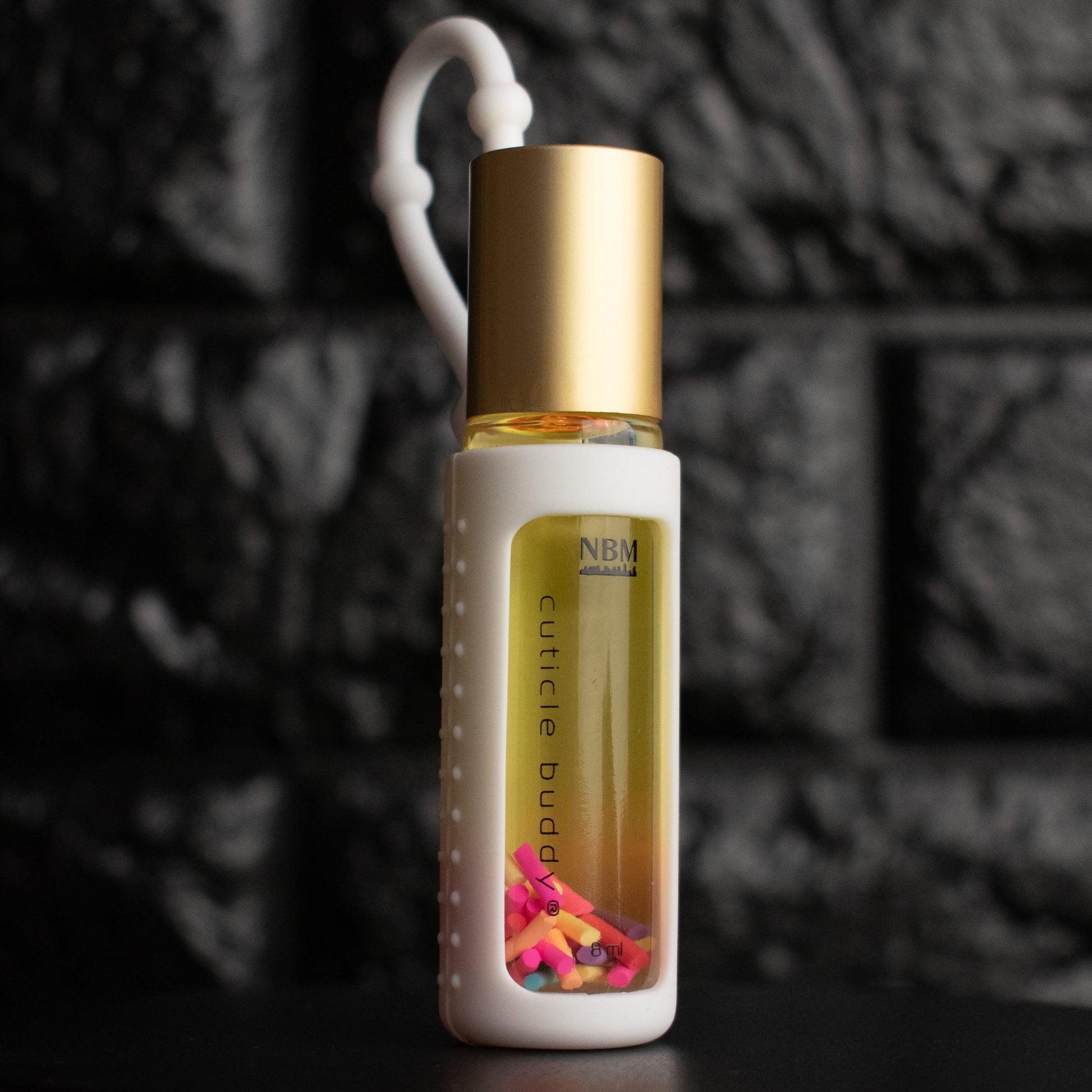 a glass bottle of oil with a gold aluminum cap in a white silicone holder on a black brick background. inside are multicolored rainbow fimo clay sprinkles