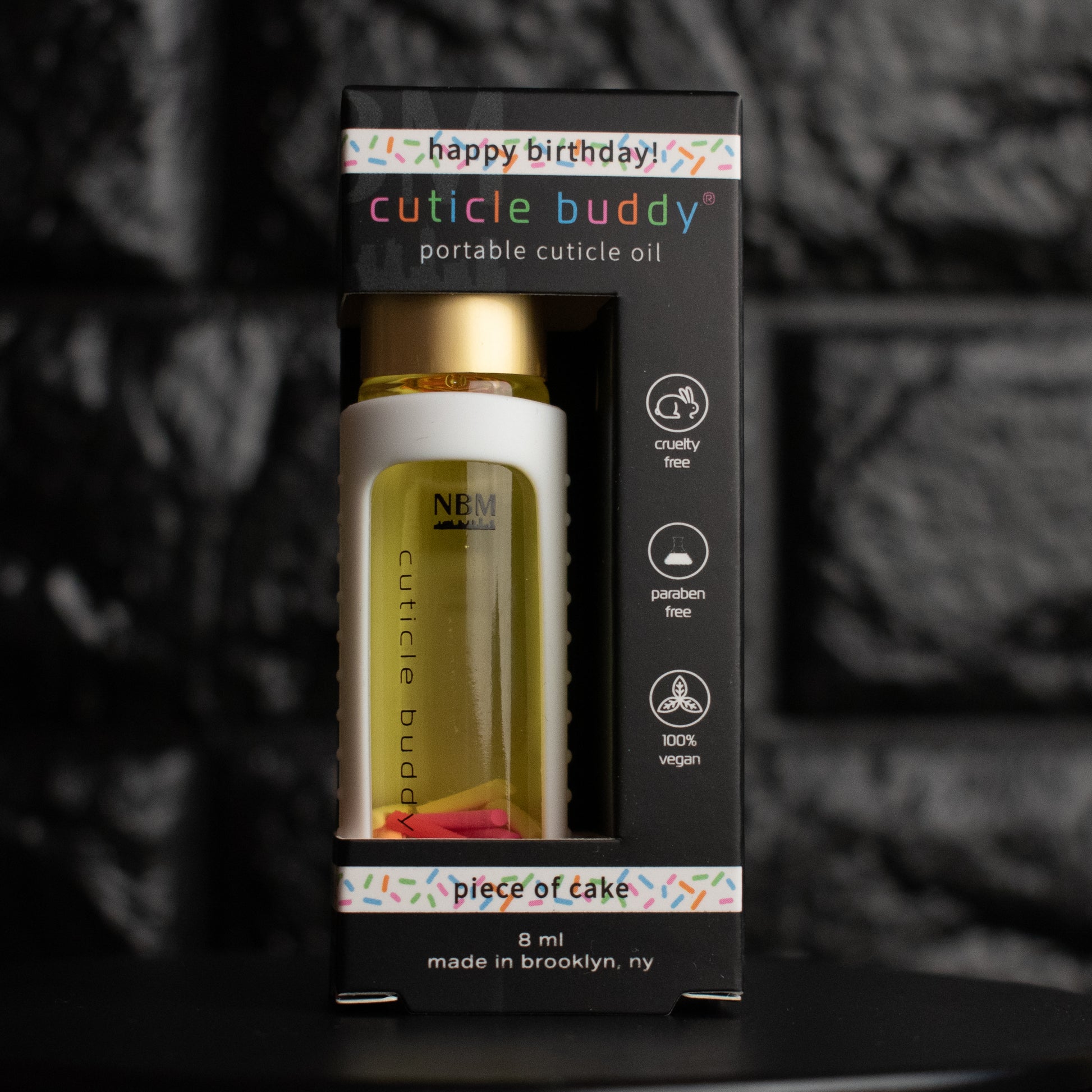 a glass bottle of oil with a gold aluminum cap in a white silicone holder in it's black packaging box on a black brick background. inside are multicolored rainbow fimo clay sprinkles