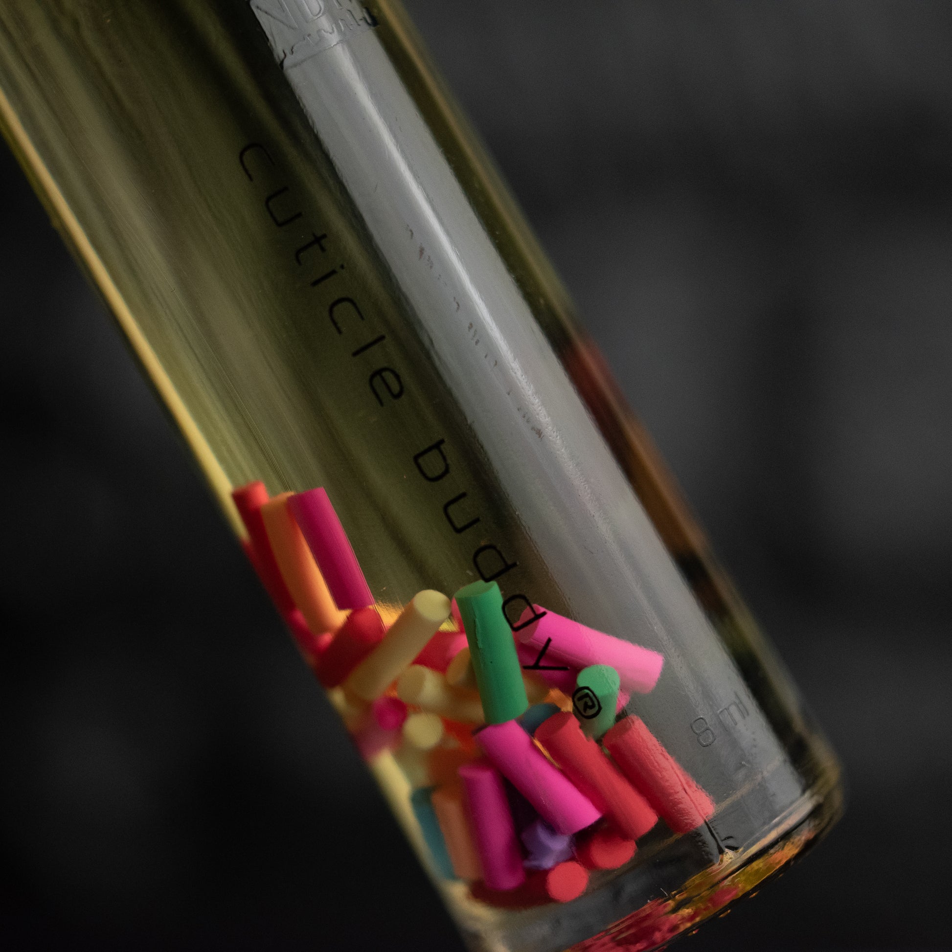 Close-up of a clear glass bottle labeled 'Cuticle Buddy' that's filled with oil and multicolored rainbow fimo sprinkles on a dark background