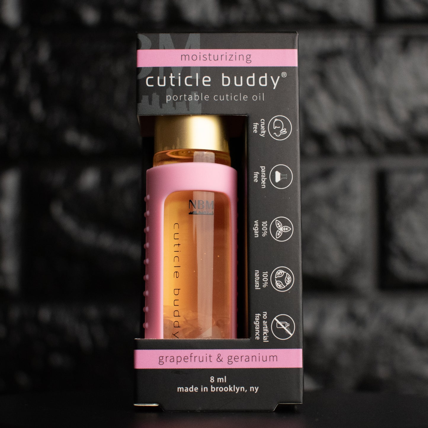 a glass bottle of oil with a gold aluminum cap in a pink silicone holder on a black brick background. inside the bottle are rose quartz stones. in its packaging box