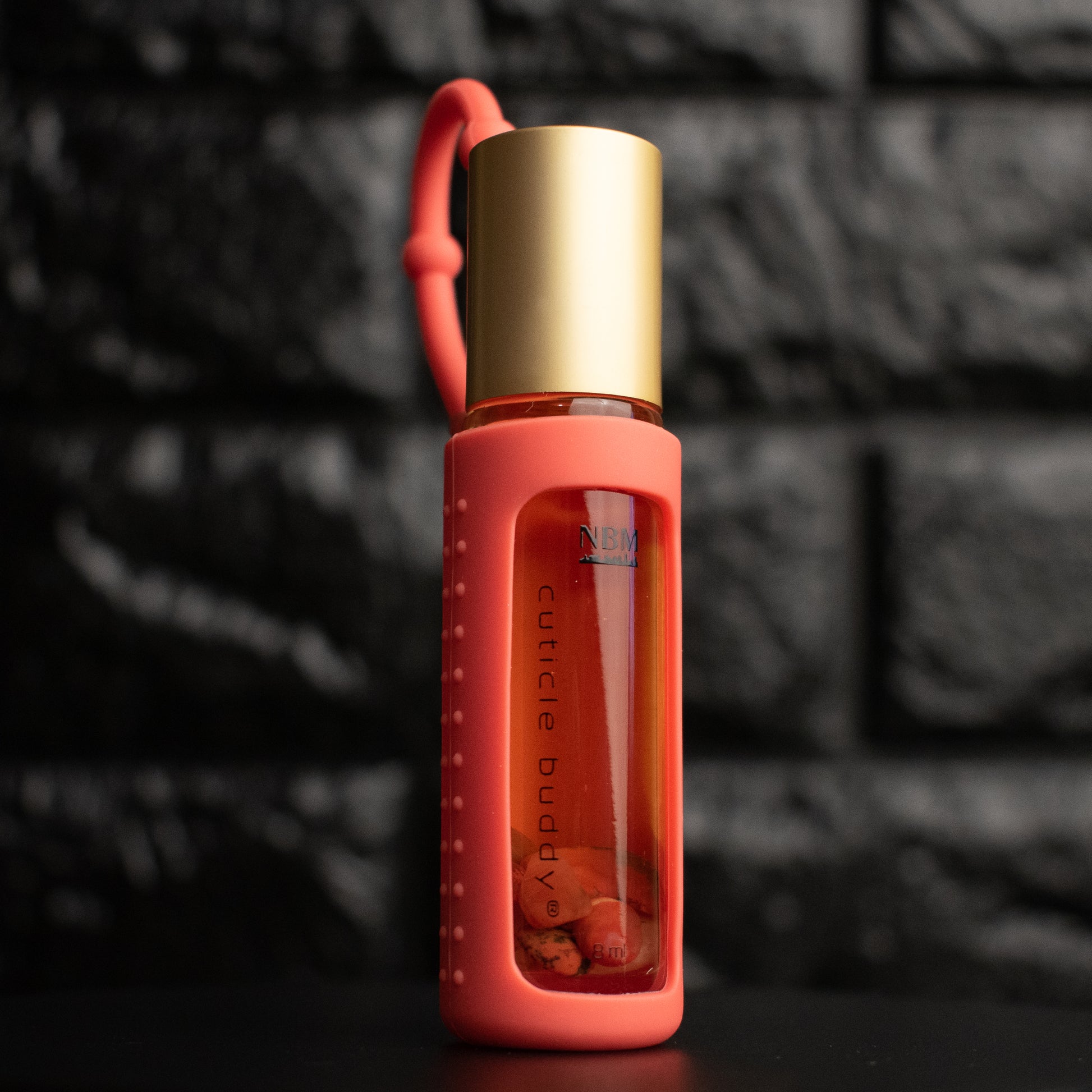 a glass bottle of oil with a gold aluminum cap in an orangesilicone holder on a black brick background. inside the bottle are red agate stones