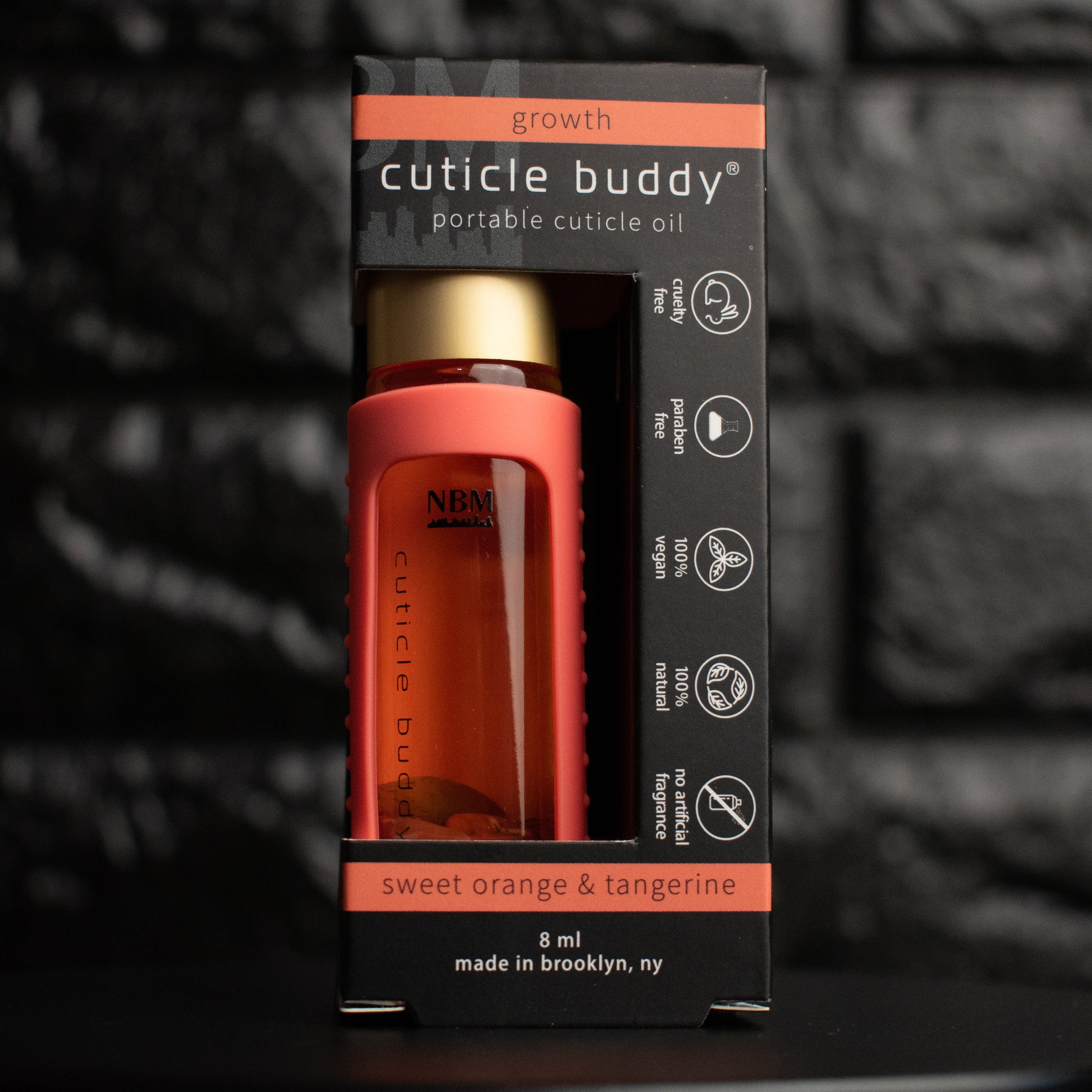 a glass bottle of oil with a gold aluminum cap in an orangesilicone holder on a black brick background. inside the bottle are red agate stones. in  its packaging box