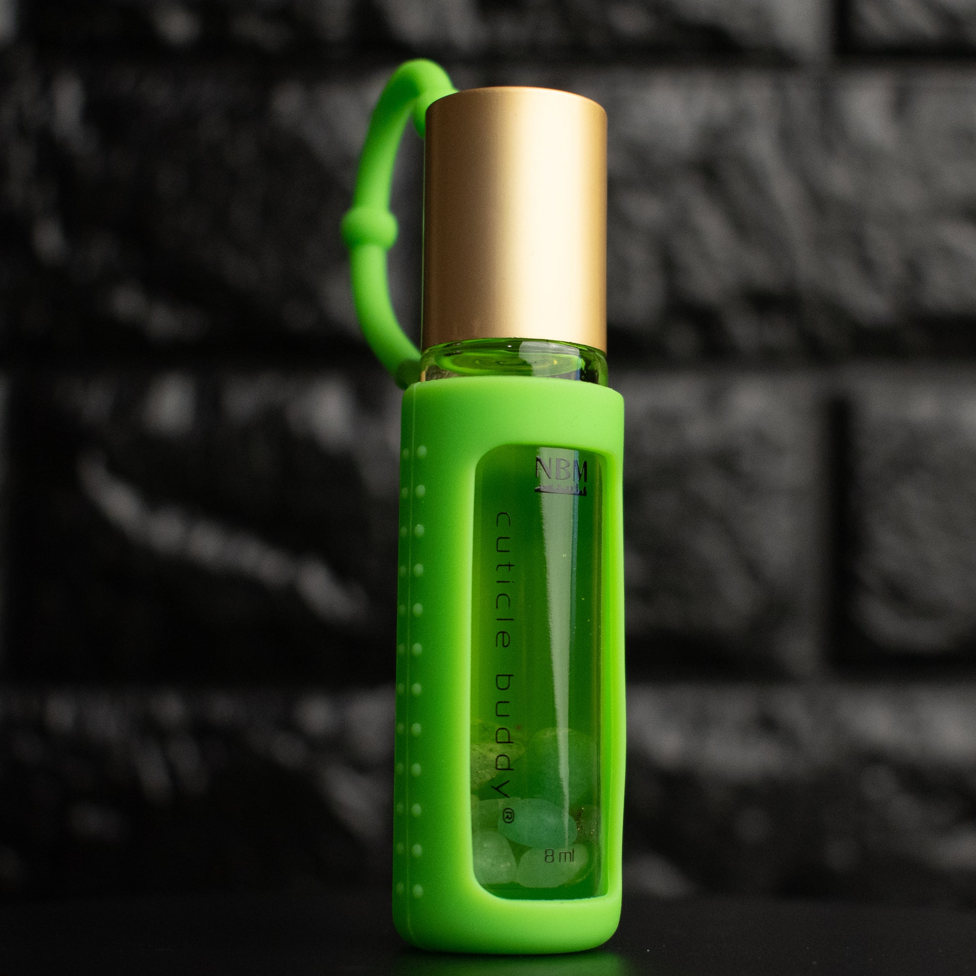 a glass bottle of oil with a gold aluminum cap in a green silicone holder on a black brick background. inside the bottle are green aventurine stones