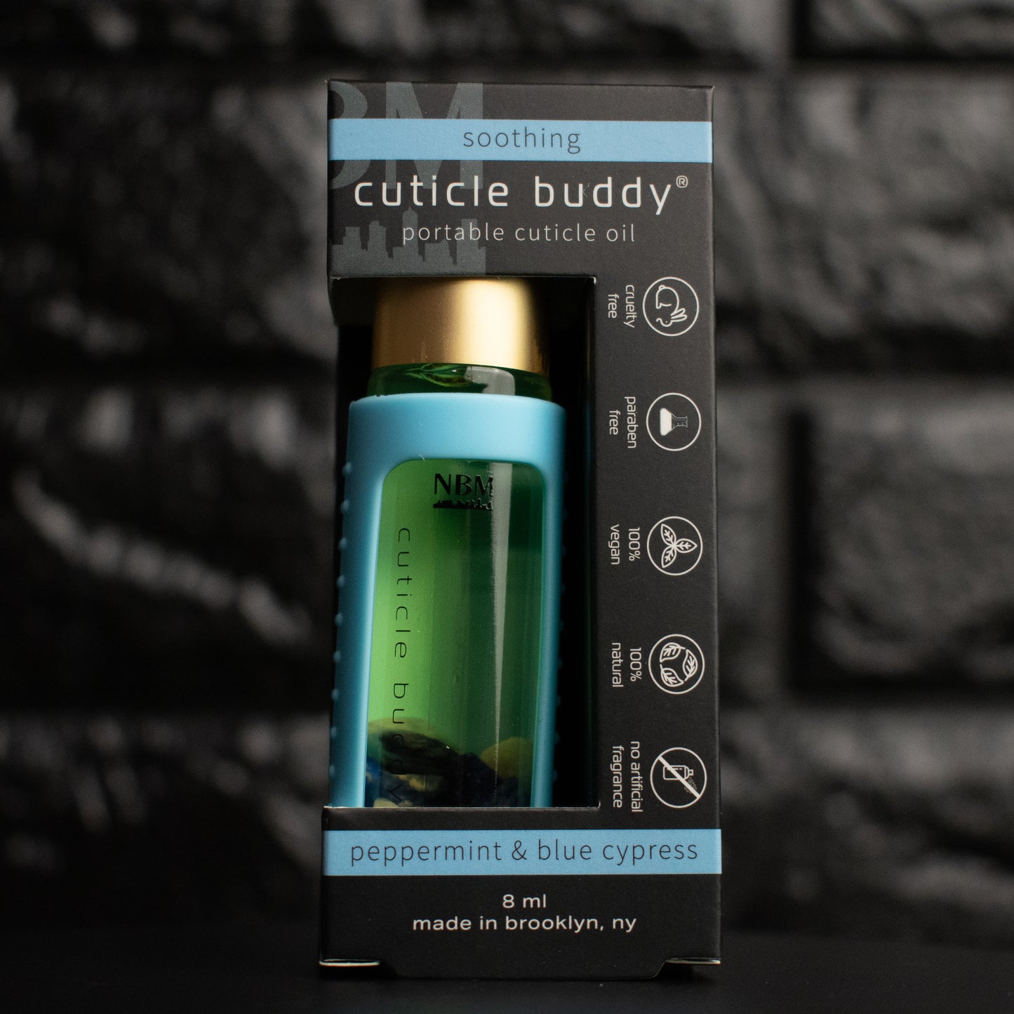 a glass bottle of oil with a gold aluminum cap in a blue silicone holder on a black brick background. inside the bottle are blue sodalite stones. in its packaging box