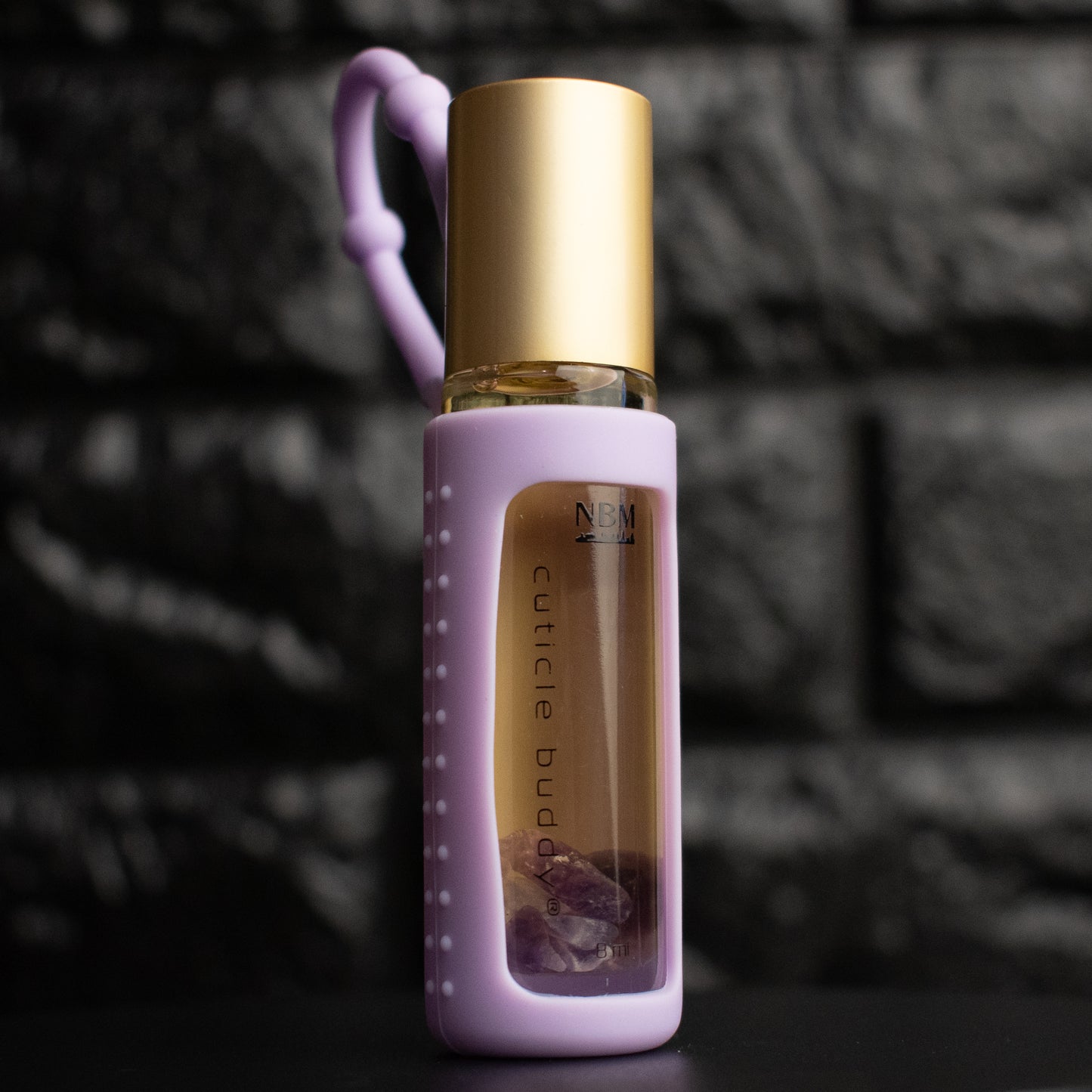 a glass bottle of oil with a gold aluminum cap in a purple silicone holder on a black brick background. inside the bottle are purple amethyst stones