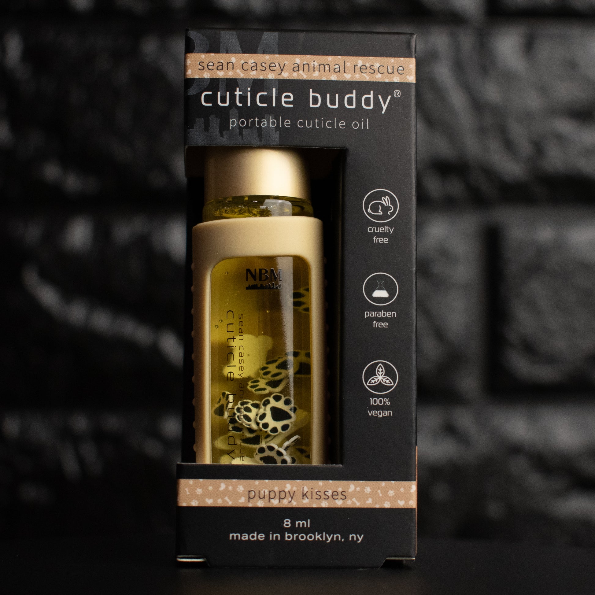 a glass bottle of oil with a gold aluminum cap in a beige silicone holder on a black brick background. inside the bottle are paw print and bone fimo clay slices. bottle is in its packaging box