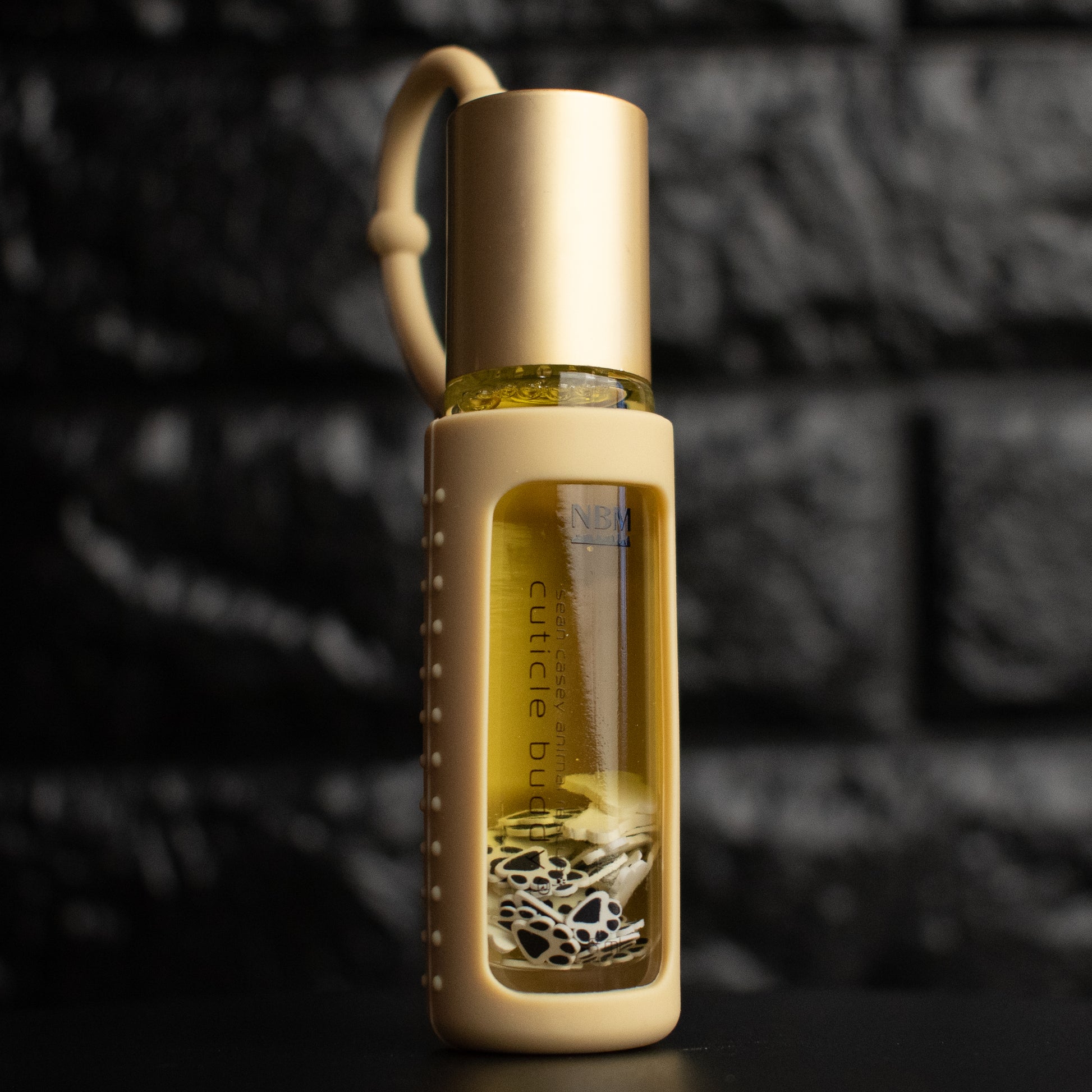 a glass bottle of oil with a gold aluminum cap in a beige silicone holder on a black brick background. inside the bottle are paw print and bone fimo clay slices