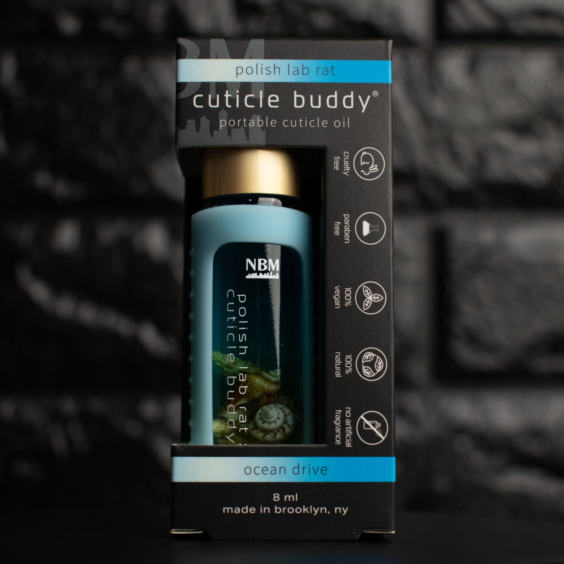 glass bottle of oil with a gold aluminum cap in a jelly blue silicone holder on a black brick background. inside the bottle are faux seashells. inside its packaging box