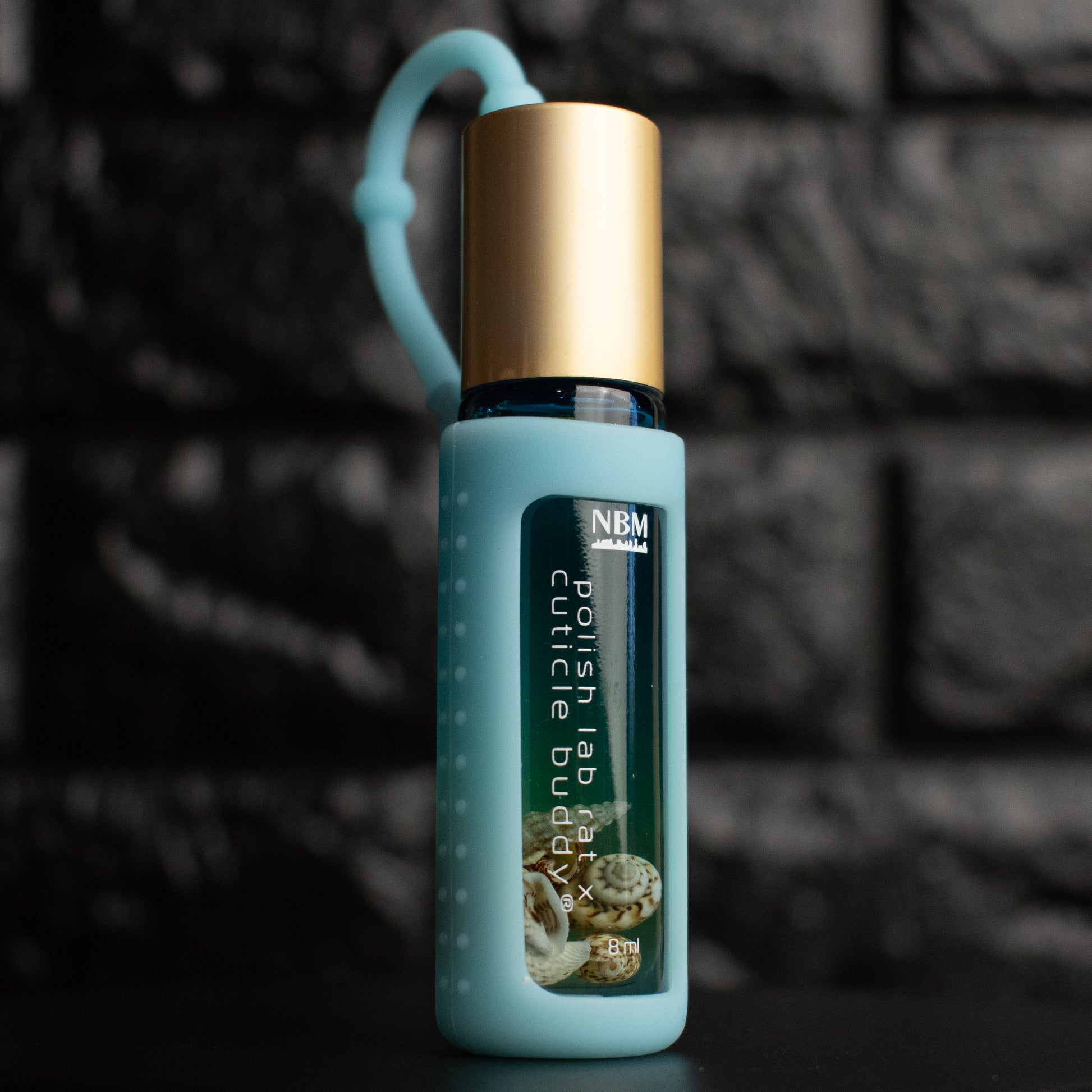 glass bottle of oil with a gold aluminum cap in a jelly blue silicone holder on a black brick background. inside the bottle are faux seashells