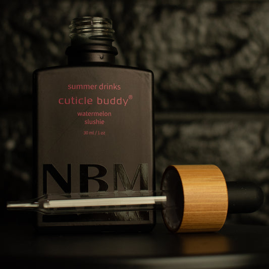 matte Black bottle with a removed wooden dropper labeled summer drinks cuticle buddy watermelon slushie 30ml/1oz a big NBM logo on the bottom featuring a city skyline. on a dark background