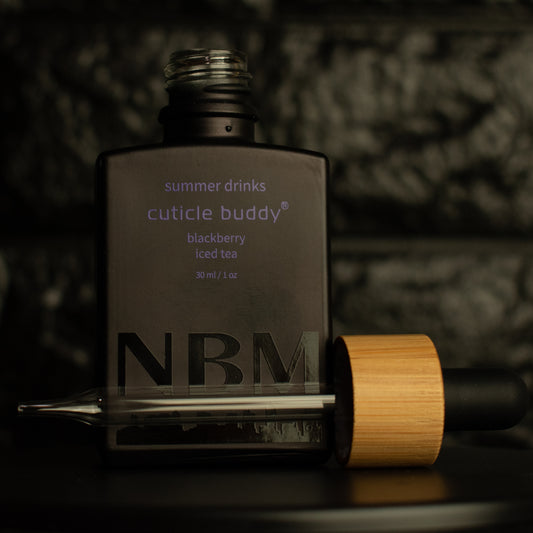 matte Black bottle with a removed wooden dropper labeled summer drinks cuticle buddy blackberry iced tea 30ml/1oz a big NBM logo on the bottom featuring a city skyline. on a dark background