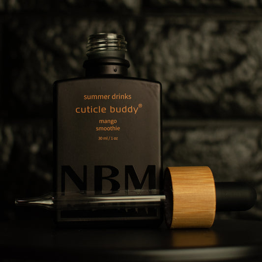 matte Black bottle with a removed wooden dropper labeled summer drinks cuticle buddy mango smoothie 30ml/1oz a big NBM logo on the bottom featuring a city skyline. on a dark background