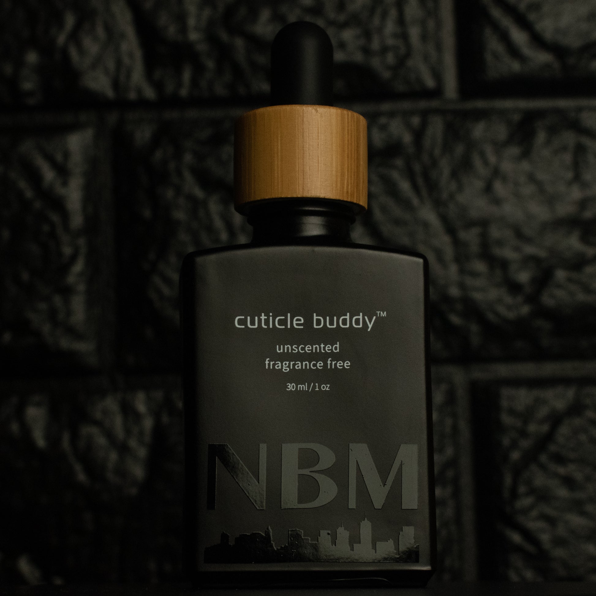 matte Black bottle with wooden dropper labeled cuticle buddy unscented fragrance free 30ml/1oz with a big NBM logo on the bottom featuring a city skyline, on  a dark background