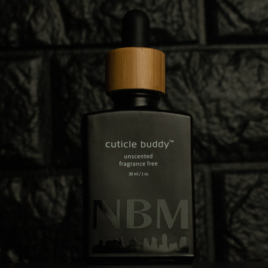 matte Black bottle with wooden dropper labeled cuticle buddy unscented fragrance free 30ml/1oz with a big NBM logo on the bottom featuring a city skyline, on  a dark background
