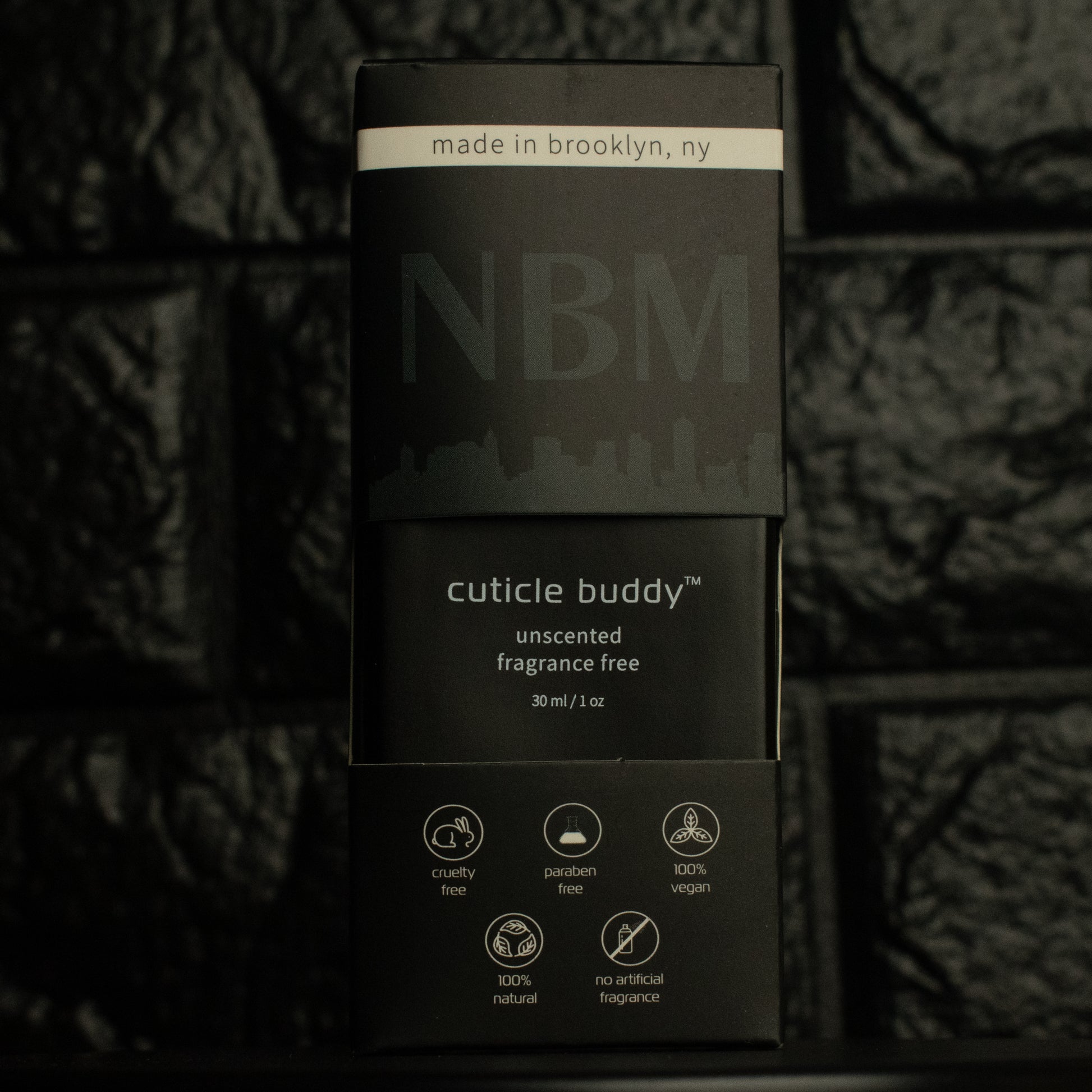 matte Black bottle with wooden dropper labeled cuticle buddy unscented fragrance free 30ml/1oz with a big NBM logo on the bottom featuring a city skyline, on  a dark background, in its packaging box