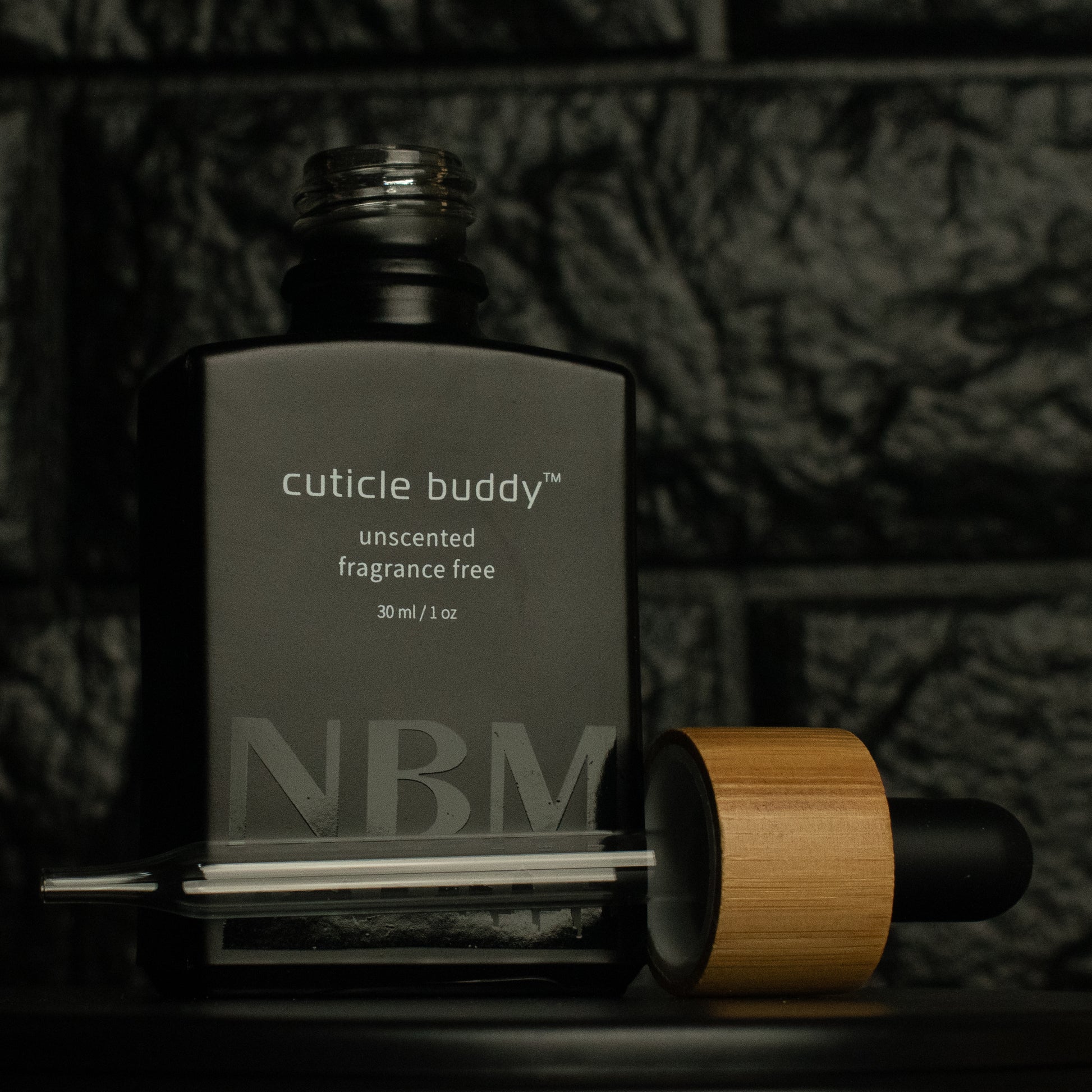 matte Black bottle with a removed wooden dropper labeled cuticle buddy unscented fragrance free 30ml/1oz with a big NBM logo on the bottom featuring a city skyline, on  a dark background