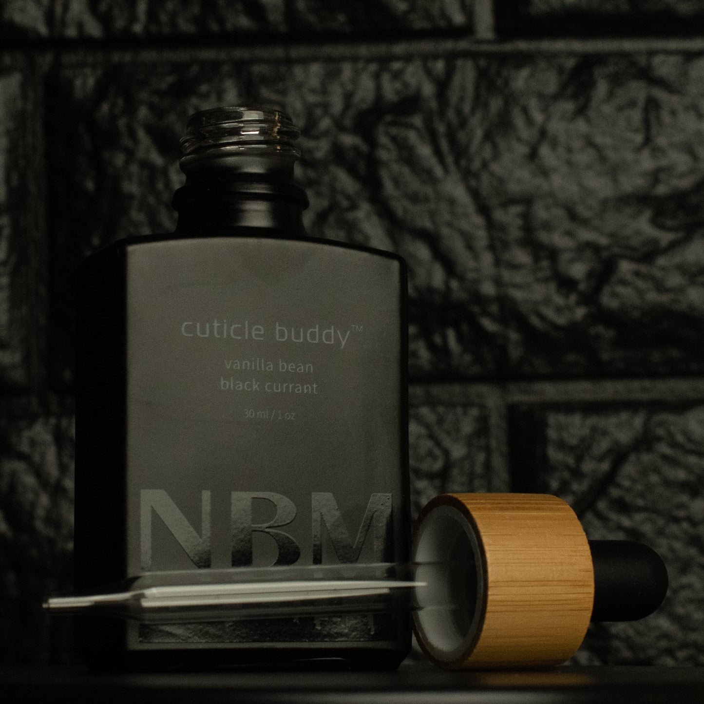 matte Black bottle with a removed wooden dropper labeled cuticle buddy vanilla bean black currant30ml/1oz with a big NBM logo on the bottom featuring a city skyline, on  a dark background