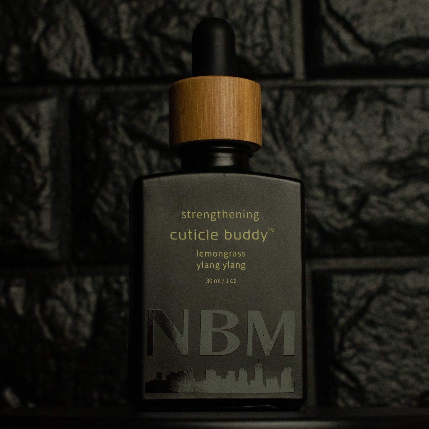matte Black bottle with wooden dropper labeled strengthening cuticle buddy lemongrass & ylang ylang 30ml/1oz with a big NBM logo on the bottom featuring a city skyline, on a dark background