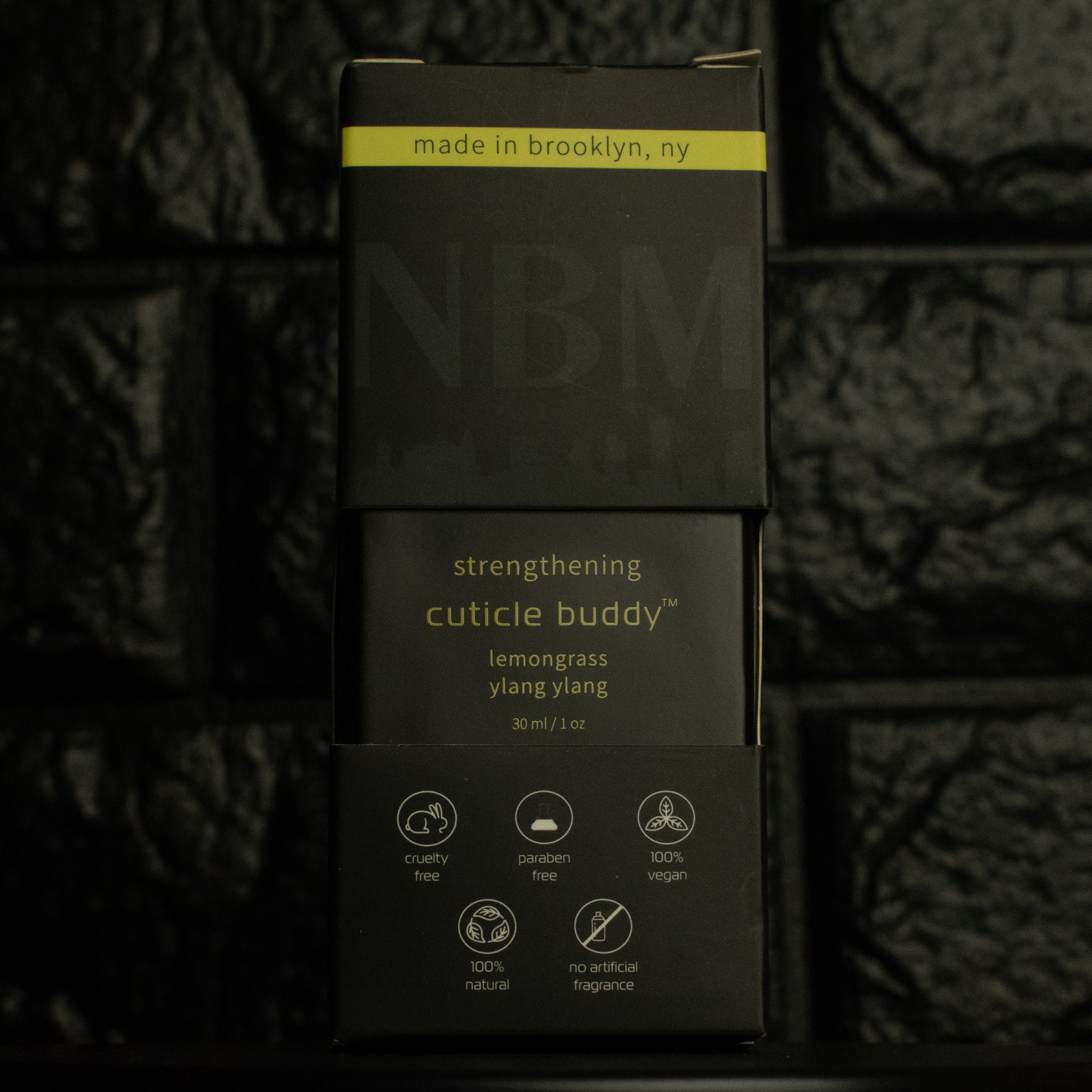 matte Black bottle with wooden dropper labeled strengthening cuticle buddy lemongrass & ylang ylang 30ml/1oz, in its packaging box, on a dark background.