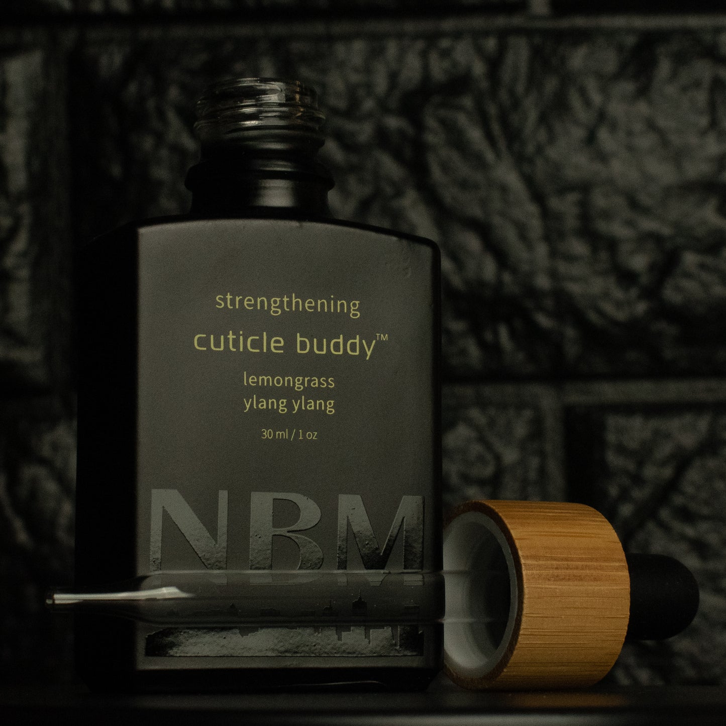 matte Black bottle with a removed wooden dropper labeled strengthening cuticle buddy lemongrass & ylang ylang 30ml/1oz with a big NBM logo on the bottom featuring a city skyline, on a dark background