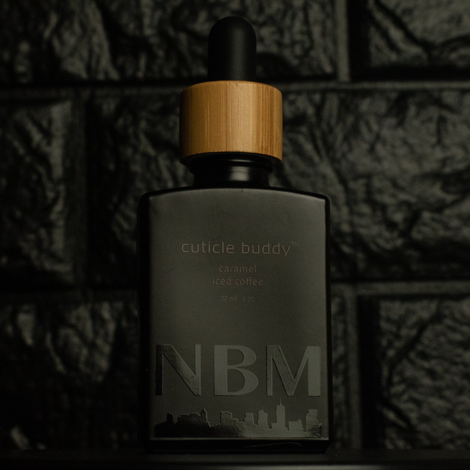 matte Black bottle with wooden dropper labeled cuticle buddy caramel iced coffee 30ml/1oz with a big NBM logo on the bottom featuring a city skyline, on  a dark background