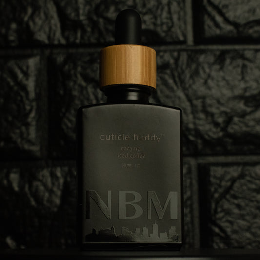 matte Black bottle with wooden dropper labeled cuticle buddy caramel iced coffee 30ml/1oz with a big NBM logo on the bottom featuring a city skyline, on  a dark background