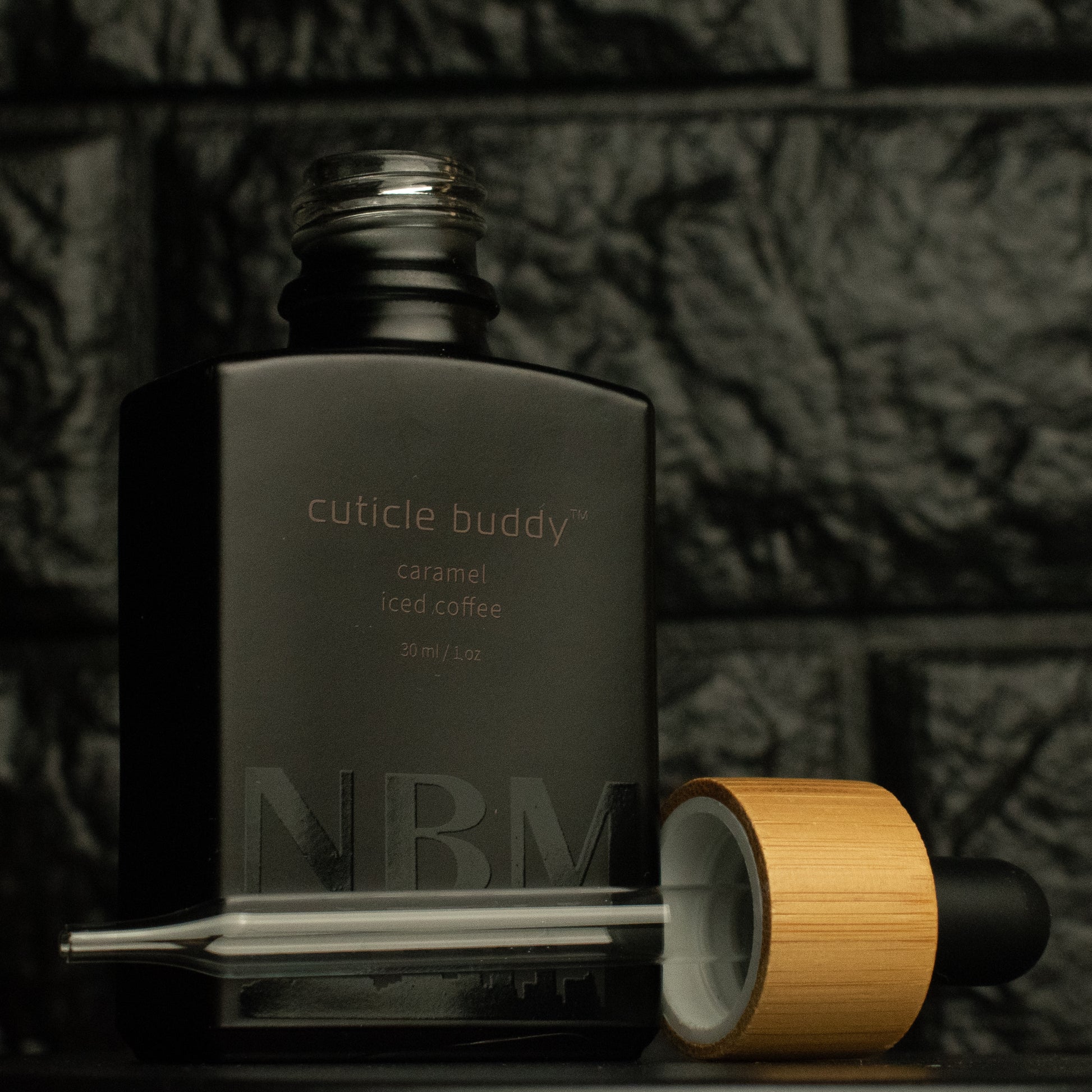 matte Black bottle with a removed wooden dropper labeled cuticle buddy caramel iced coffee 30ml/1oz with a big NBM logo on the bottom featuring a city skyline, on  a dark background