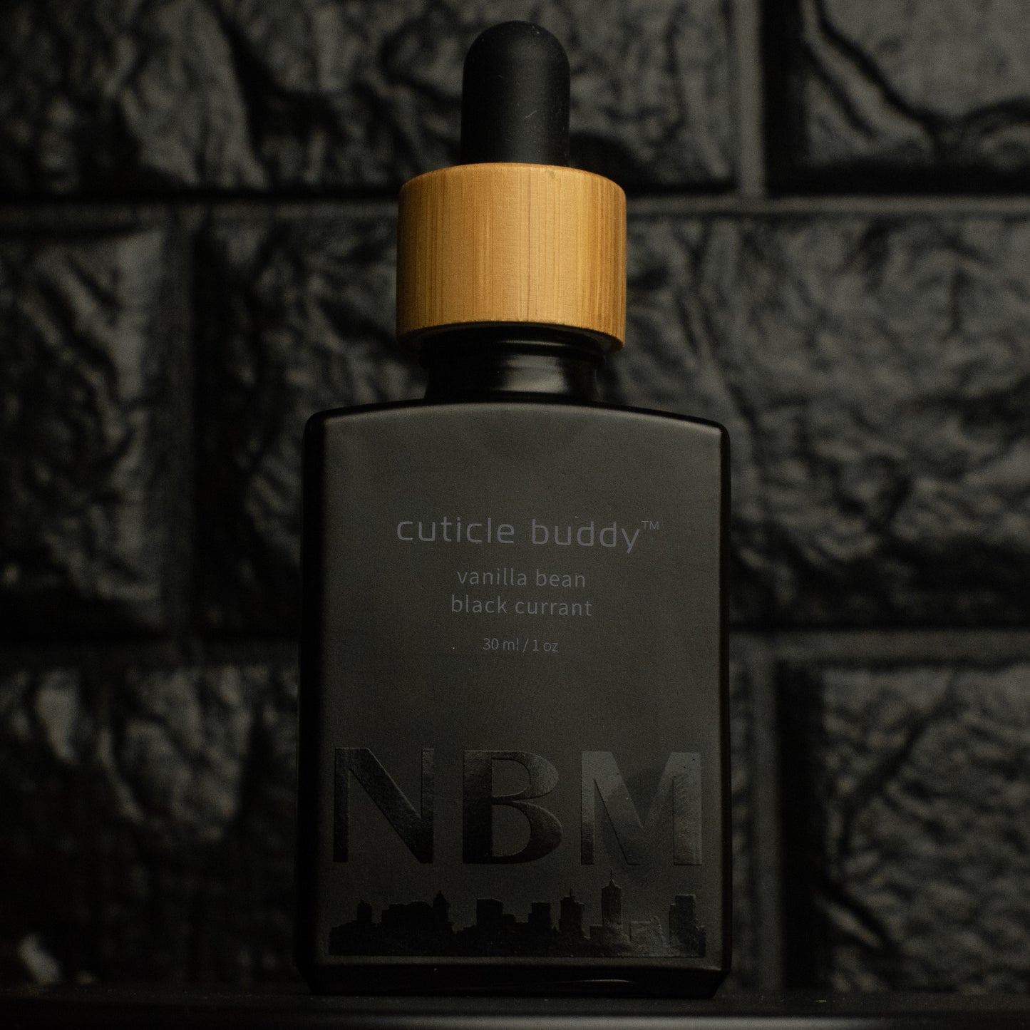 matte Black bottle with wooden dropper labeled cuticle buddy vanilla bean black currant30ml/1oz with a big NBM logo on the bottom featuring a city skyline, on  a dark background