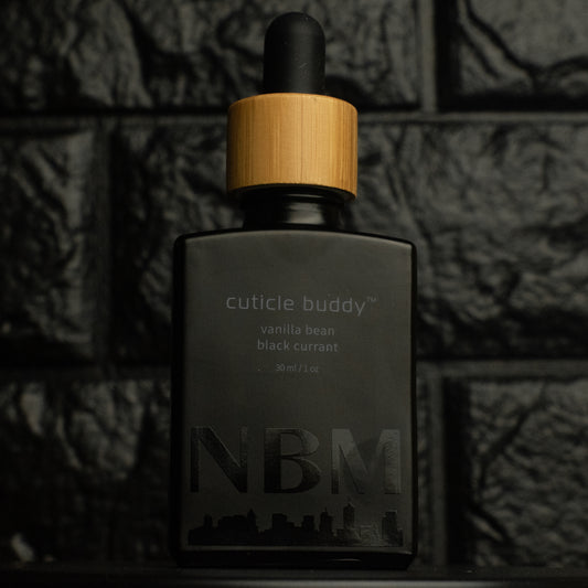 matte Black bottle with wooden dropper labeled cuticle buddy vanilla bean black currant30ml/1oz with a big NBM logo on the bottom featuring a city skyline, on  a dark background