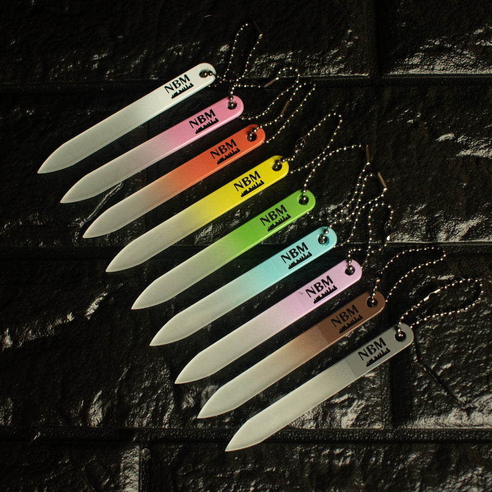 a collection of 9 different portable mini glass nail files branded with the NBM logo in white, pink, orange, yellow, green, blue, purple, brown and gray, each with a black ball chain attached on a black brick background