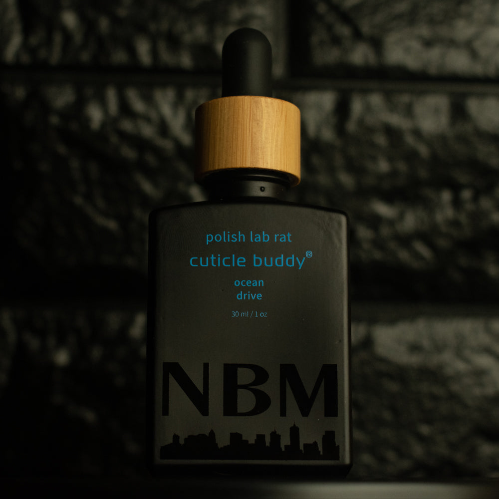 matte Black bottle with wooden dropper labeled polish lab rat cuticle buddy ocean drive 30ml/1oz with a big NBM logo on the bottom featuring a city skyline, on  a dark background