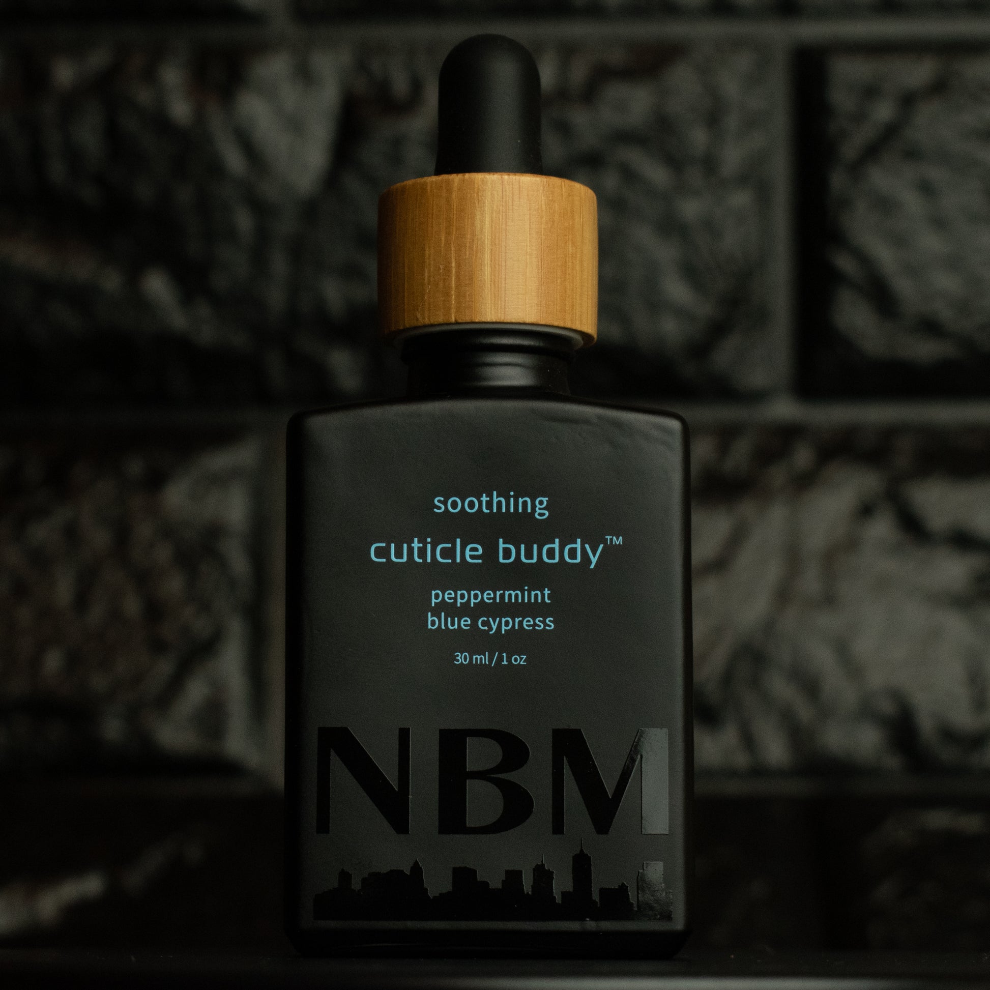 matte Black bottle with wooden dropper labeled soothing cuticle buddy peppermint blue cypress 30ml/1oz with a big NBM logo on the bottom featuring a city skyline, on  a dark background