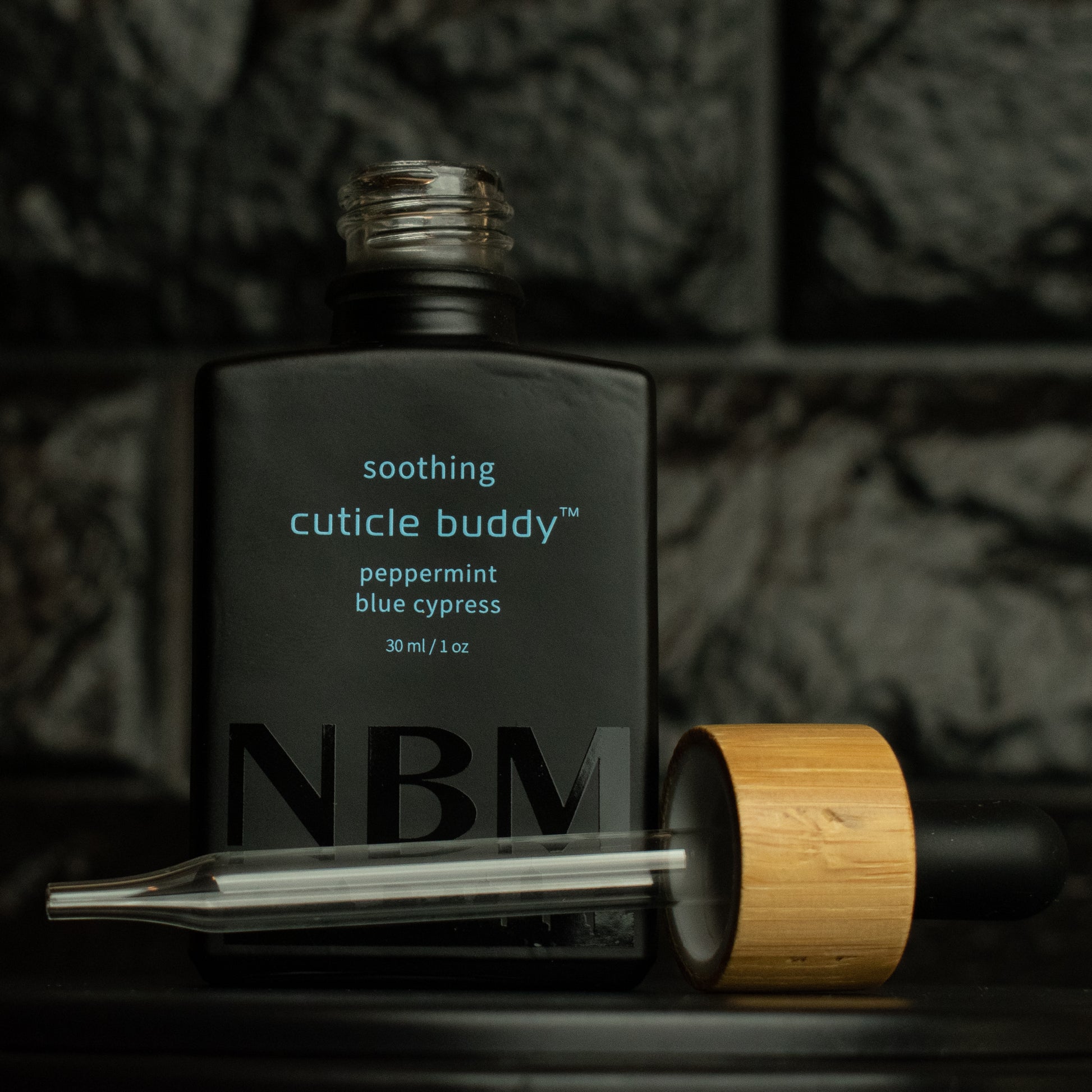 matte Black bottle with a removed wooden dropper labeled soothing cuticle buddy peppermint blue cypress 30ml/1oz with a big NBM logo on the bottom featuring a city skyline, on  a dark background