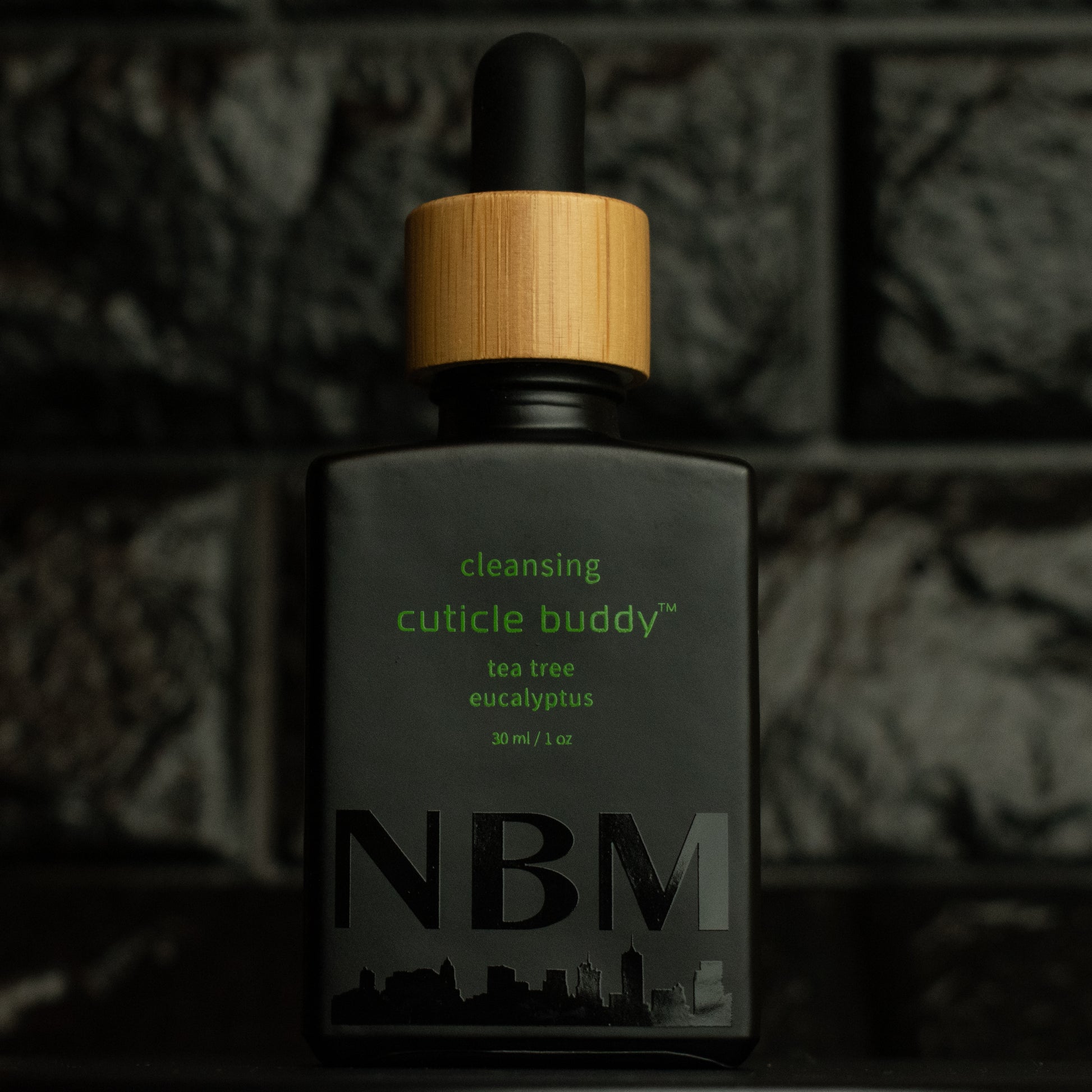 matte Black bottle with wooden dropper labeled cleansing cuticle buddy tea tree eucalyptus 30ml/1oz with a big NBM logo on the bottom featuring a city skyline, on  a dark background