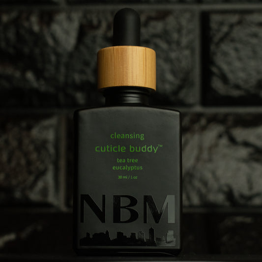 matte Black bottle with wooden dropper labeled cleansing cuticle buddy tea tree eucalyptus 30ml/1oz with a big NBM logo on the bottom featuring a city skyline, on  a dark background