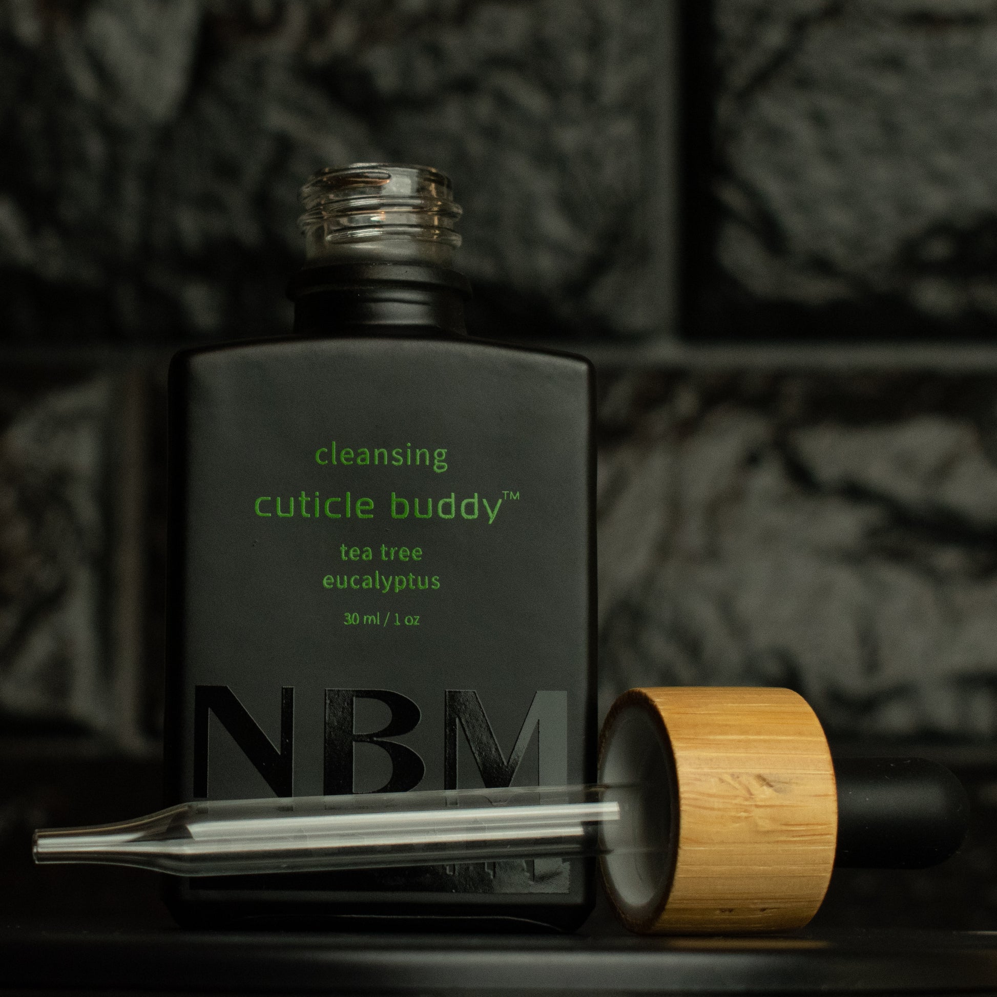 matte Black bottle with a removed wooden dropper labeled cleansing cuticle buddy tea tree eucalyptus 30ml/1oz with a big NBM logo on the bottom featuring a city skyline, on  a dark background