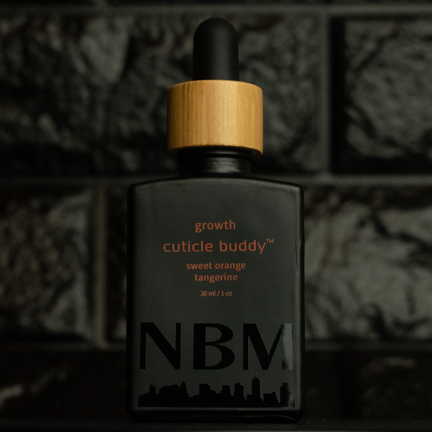 matte Black bottle with wooden dropper labeled growth cuticle buddy sweet orange tangerine 30ml/1oz with a big NBM logo on the bottom featuring a city skyline, on a dark background