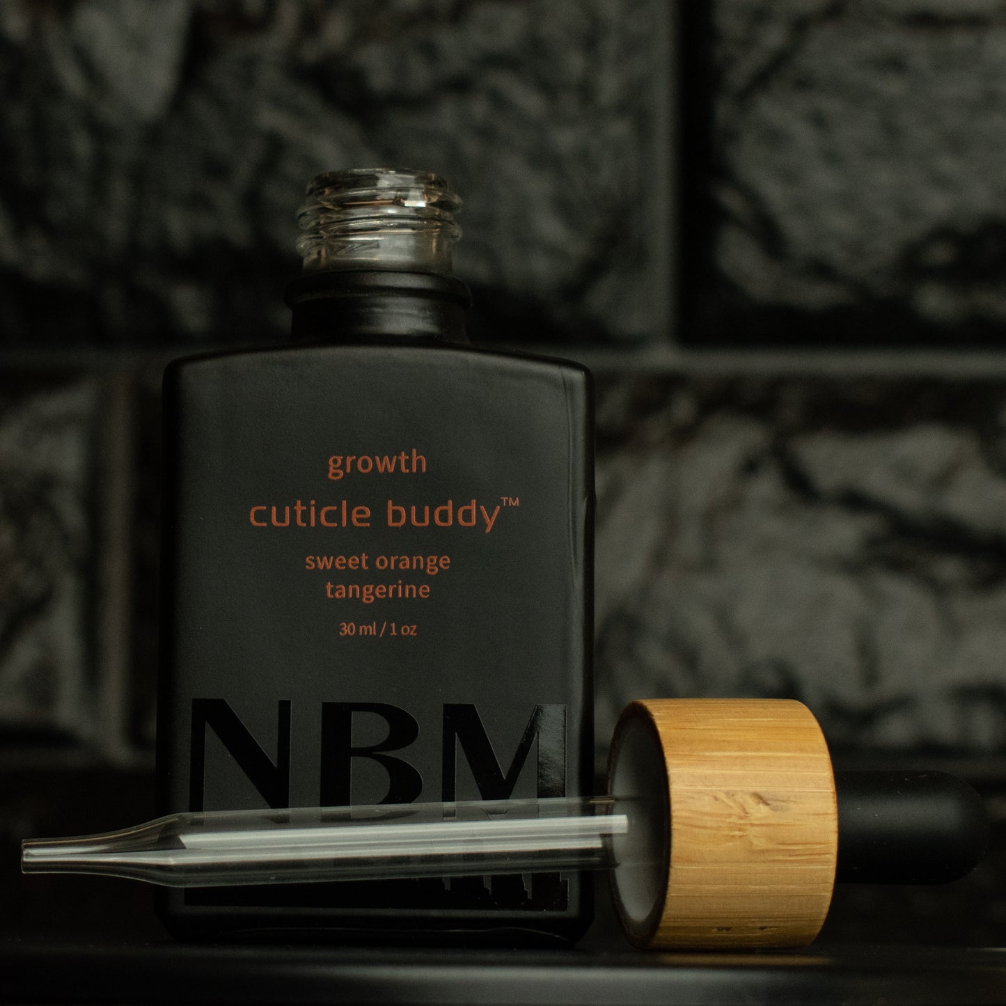 matte Black bottle with a removed wooden dropper labeled growth cuticle buddy sweet orange tangerine 30ml/1oz with a big NBM logo on the bottom featuring a city skyline, on a dark background
