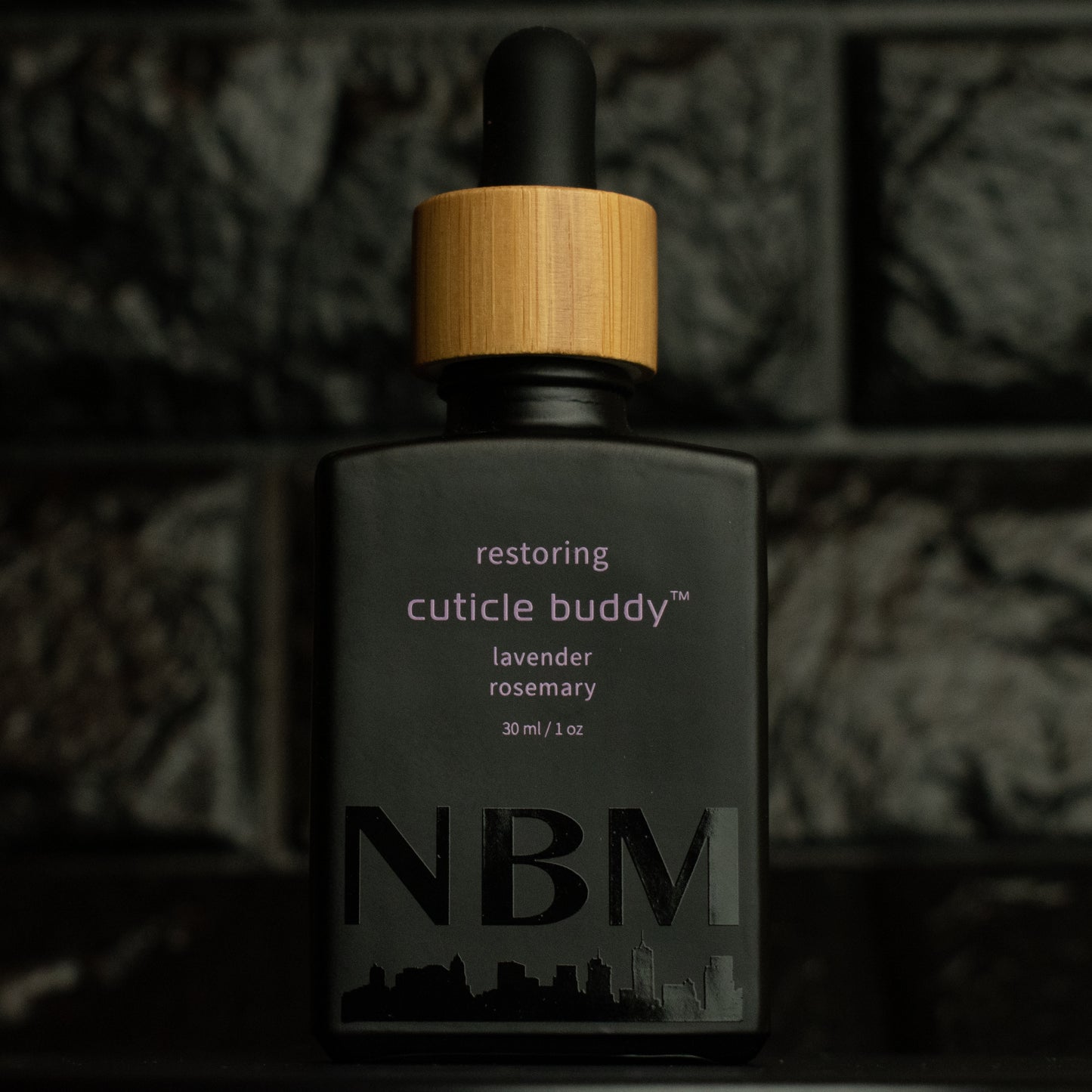matte Black bottle with wooden dropper labeled restoring cuticle buddy lavender rosemary 30ml/1oz with a big NBM logo on the bottom featuring a city skyline, on a dark background