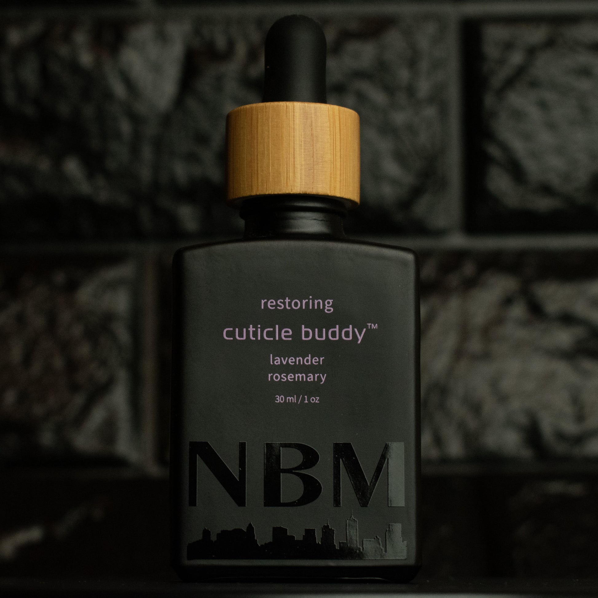 matte Black bottle with wooden dropper labeled restoring cuticle buddy lavender rosemary 30ml/1oz with a big NBM logo on the bottom featuring a city skyline, on  a dark background