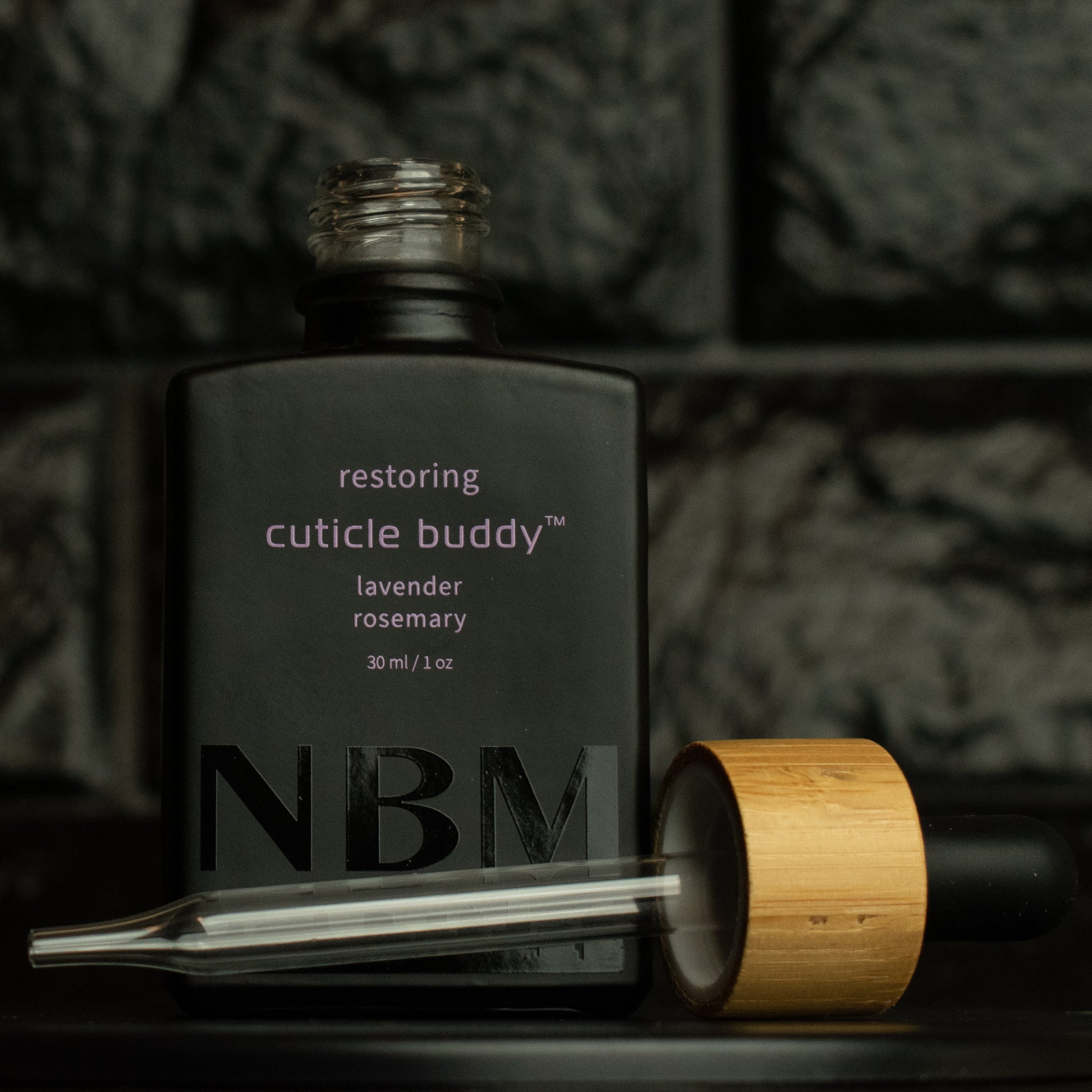 matte Black bottle with a removed wooden dropper labeled restoring cuticle buddy lavender rosemary 30ml/1oz with a big NBM logo on the bottom featuring a city skyline, on  a dark background