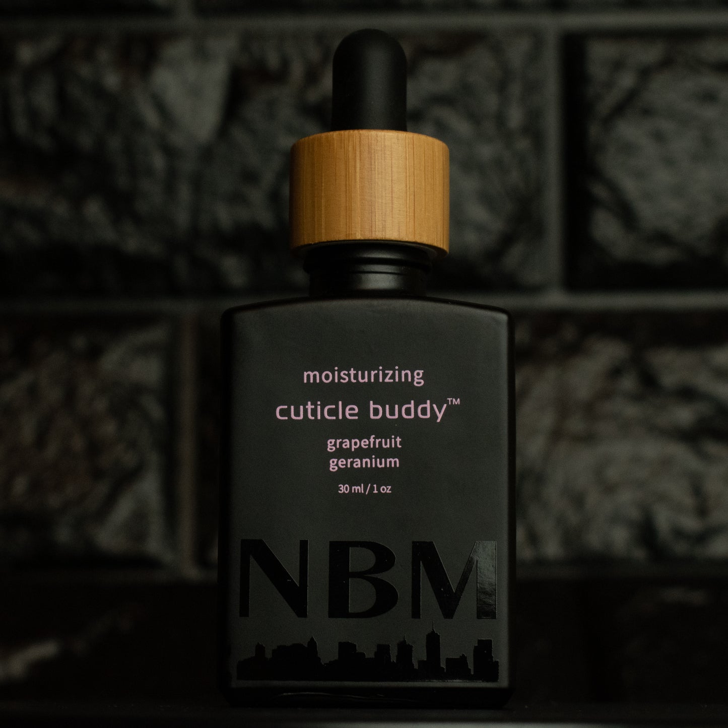 matte Black bottle with wooden dropper labeled moisturizing cuticle buddy grapefruit geranium 30ml/1oz with a big NBM logo on the bottom featuring a city skyline, on  a dark background