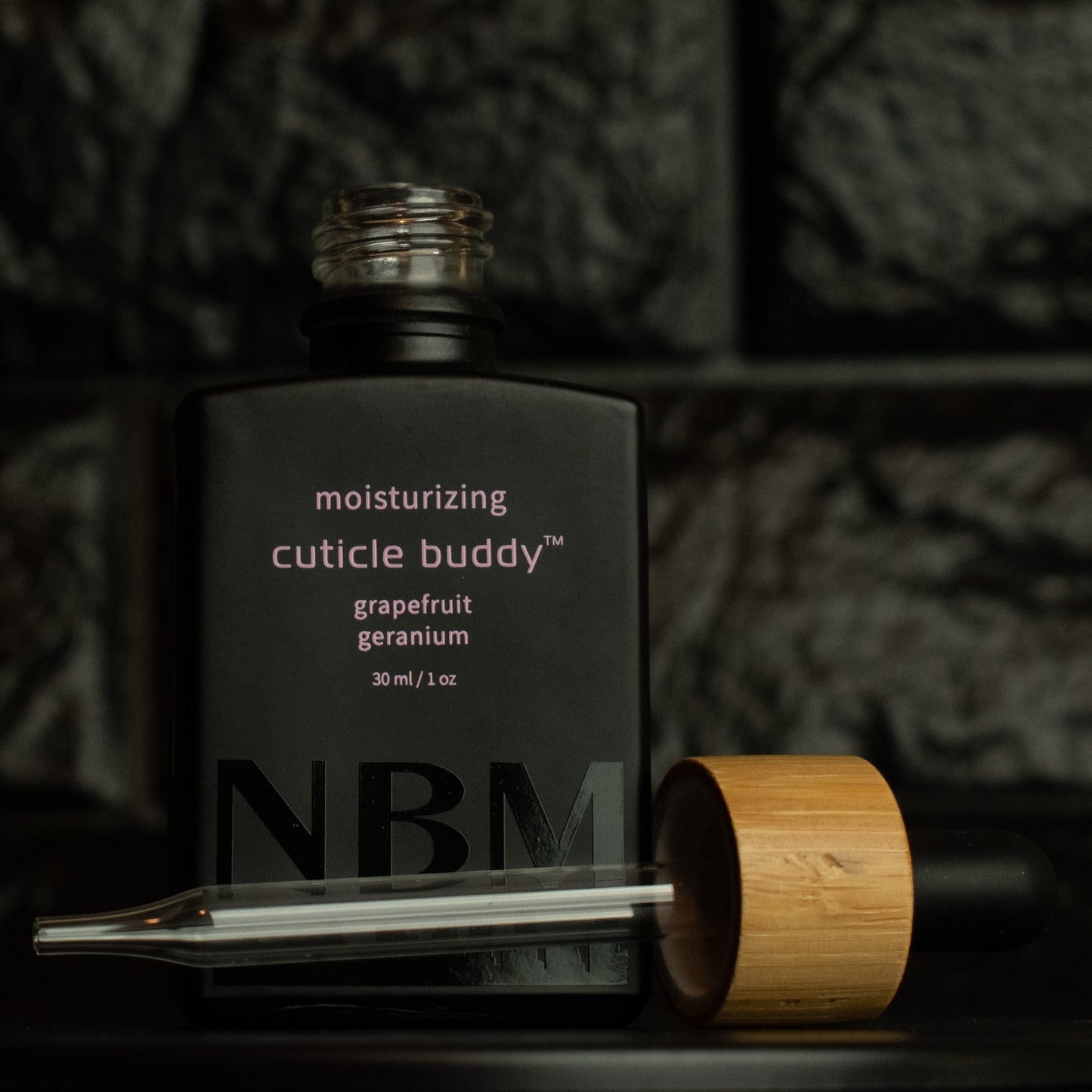 matte Black bottle with a removed wooden dropper labeled moisturizing cuticle buddy grapefruit geranium 30ml/1oz with a big NBM logo on the bottom featuring a city skyline, on  a dark background