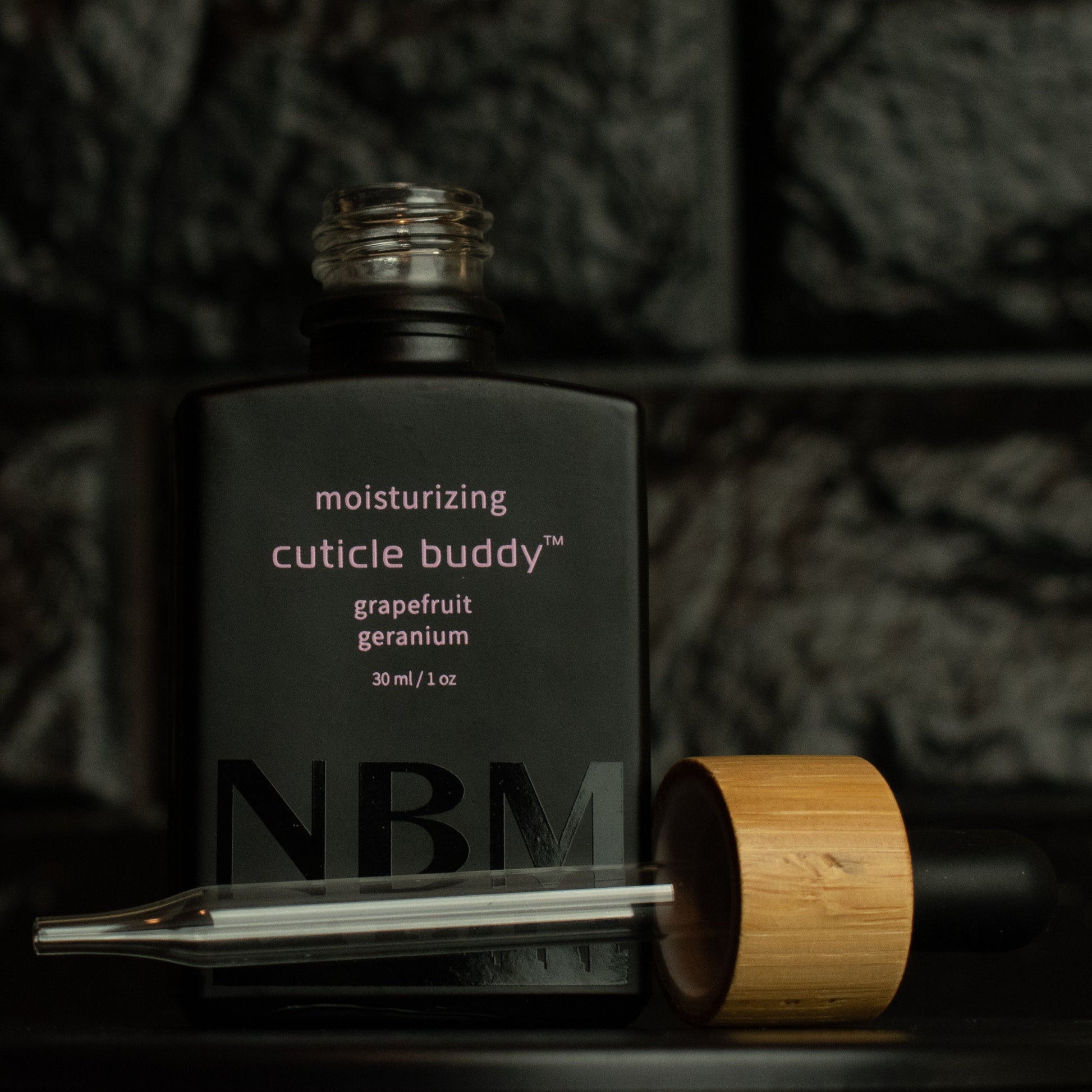 matte Black bottle with a removed wooden dropper labeled moisturizing cuticle buddy grapefruit geranium 30ml/1oz with a big NBM logo on the bottom featuring a city skyline, on  a dark background