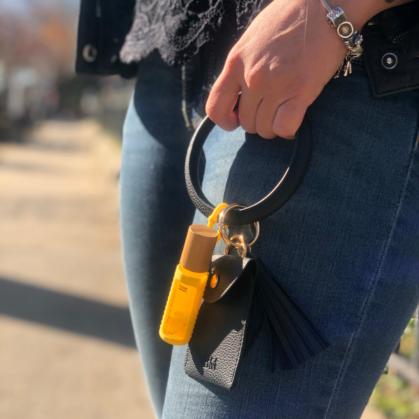 a person wearing a black lace top and dark blue jeans holding a black leather bangle wallet with a bottle of cuticle oil in a yellow silicone holder hanging off the bangle